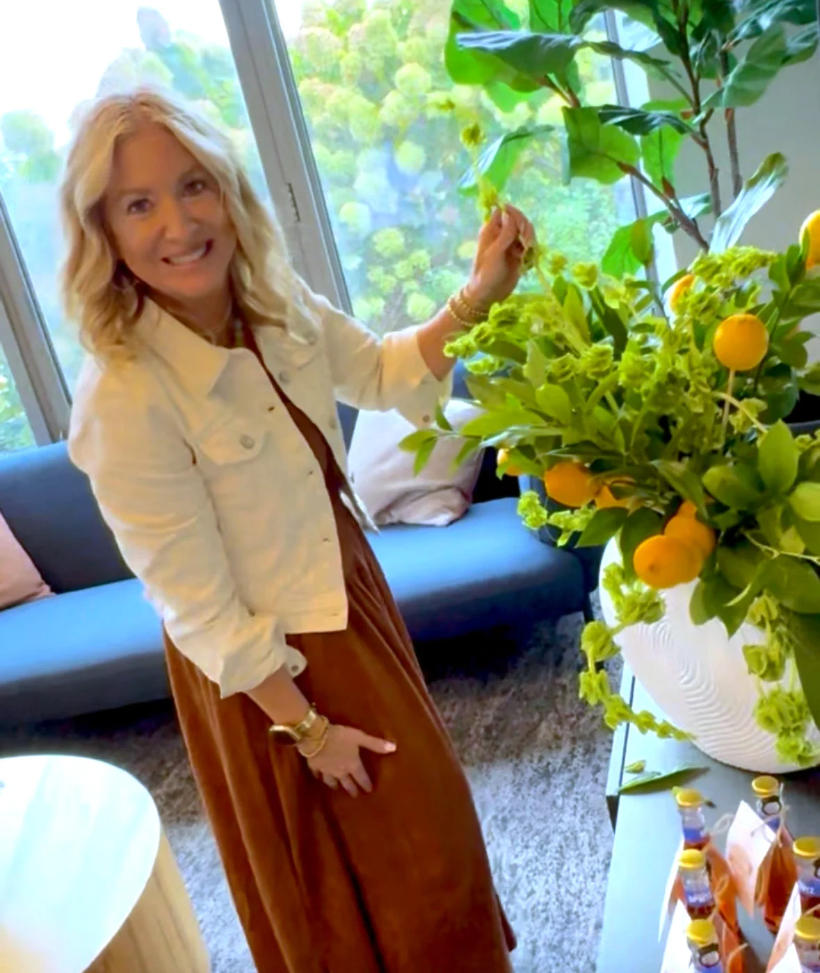 A smiling blonde woman standing next to a large, vibrant floral arrangement in a white urn. The arrangement features lush green foliage and small citrus fruits like lemons and oranges, set against a modern indoor backdrop with a blue sofa.